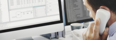 Mitarbeiter telefoniert am Arbeitsplatz vor zwei Monitoren mit CAE-Software und Tabellendaten – Symbol für COMOS Support und Administration bei A.Fasselt Engineering GmbH.