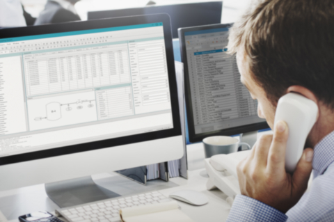Mitarbeiter telefoniert am Arbeitsplatz vor zwei Monitoren mit CAE-Software und Tabellendaten – Symbol für COMOS Support und Administration bei A.Fasselt Engineering GmbH.
